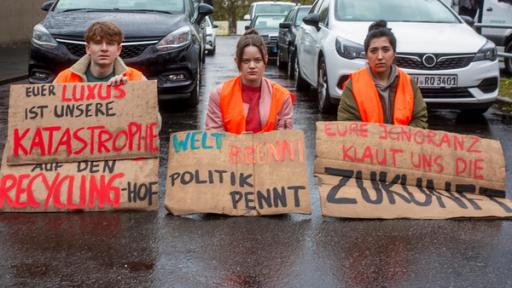 Drei Jugendliche sitzen auf einer Straße, sie tragen Warnwesten und Protestschilder