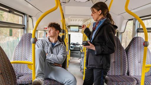 Im hinteren Teil eines Busses sitzt ein Jugendlicher im grauen Jogginganzug. Er hat seine Arme verschränkt und seinen rechten Fuß auf dem Sitz vor ihm abgestellt. Trotzig und genervt schaut er aus dem Fenster und ignoriert die Frau, die neben ihm steht. Sie trägt eine dunkle Jacke und eine blaue Bluse und schaut ihn tadelnd an. In der linken Hand hält sie zwei kleine Geräte zur Ticketkontrolle, den Zeigefinger der rechten Hand hat sie streng erhoben und zeigt in Richtung Ausgang des Busses.