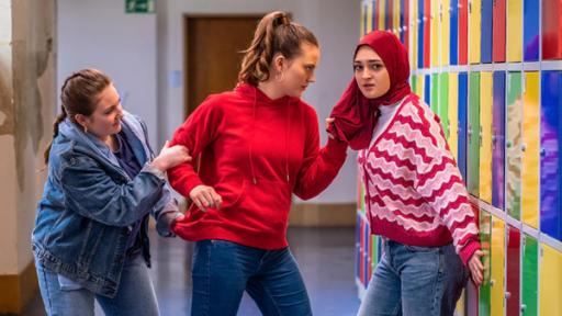 Auf einem Schulflur mit vielen bunten Schließfächern stehen drei Mädchen. Ein Mädchen trägt einen braunen Zopf und einen roten Pullover, ein anderes Mädchen trägt ein rotes Kopftuch und eine rosa-farbene Strickjacke. Das Mädchen mit Kopftuch blickt verzweifelt in die Kamera, während das Mädchen im roten Pullover aggressiv an ihrem Kopftuch zieht. 